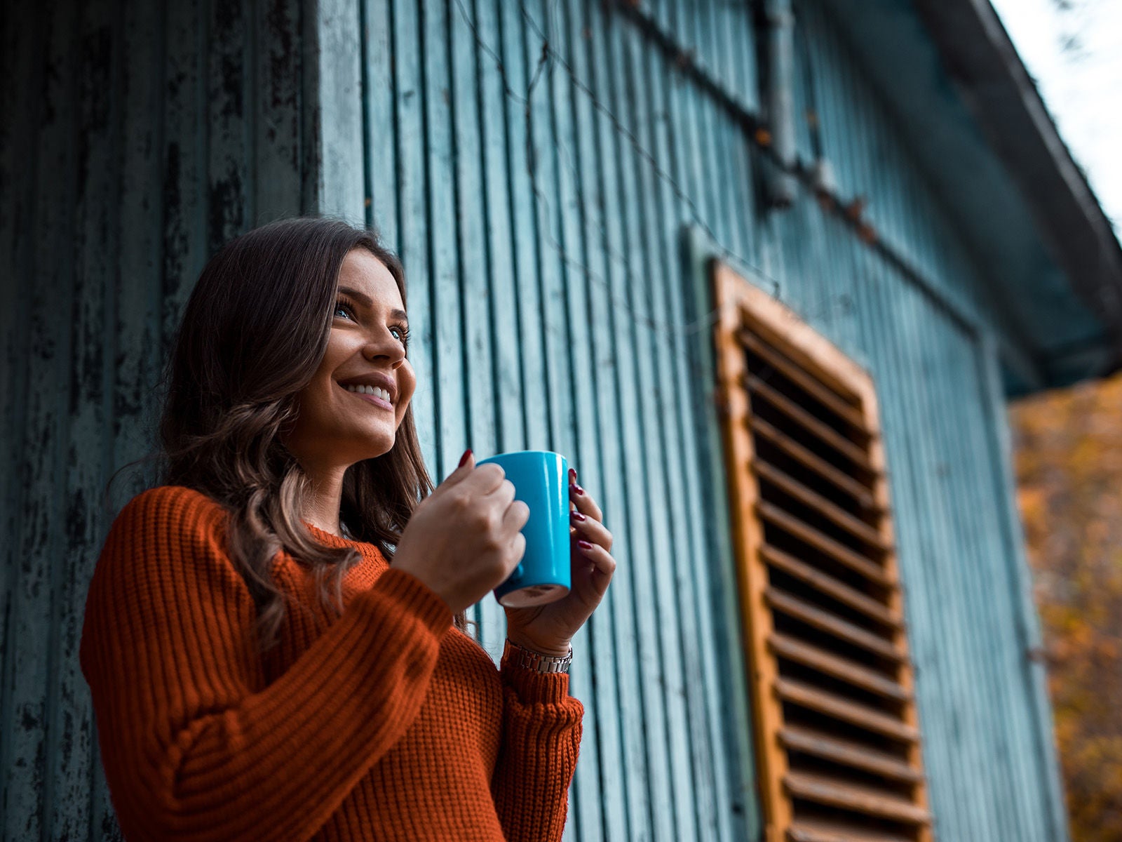 Femme souriante et décontractée tenant une tasse de café, debout à côté d’une maison en bois bleue.