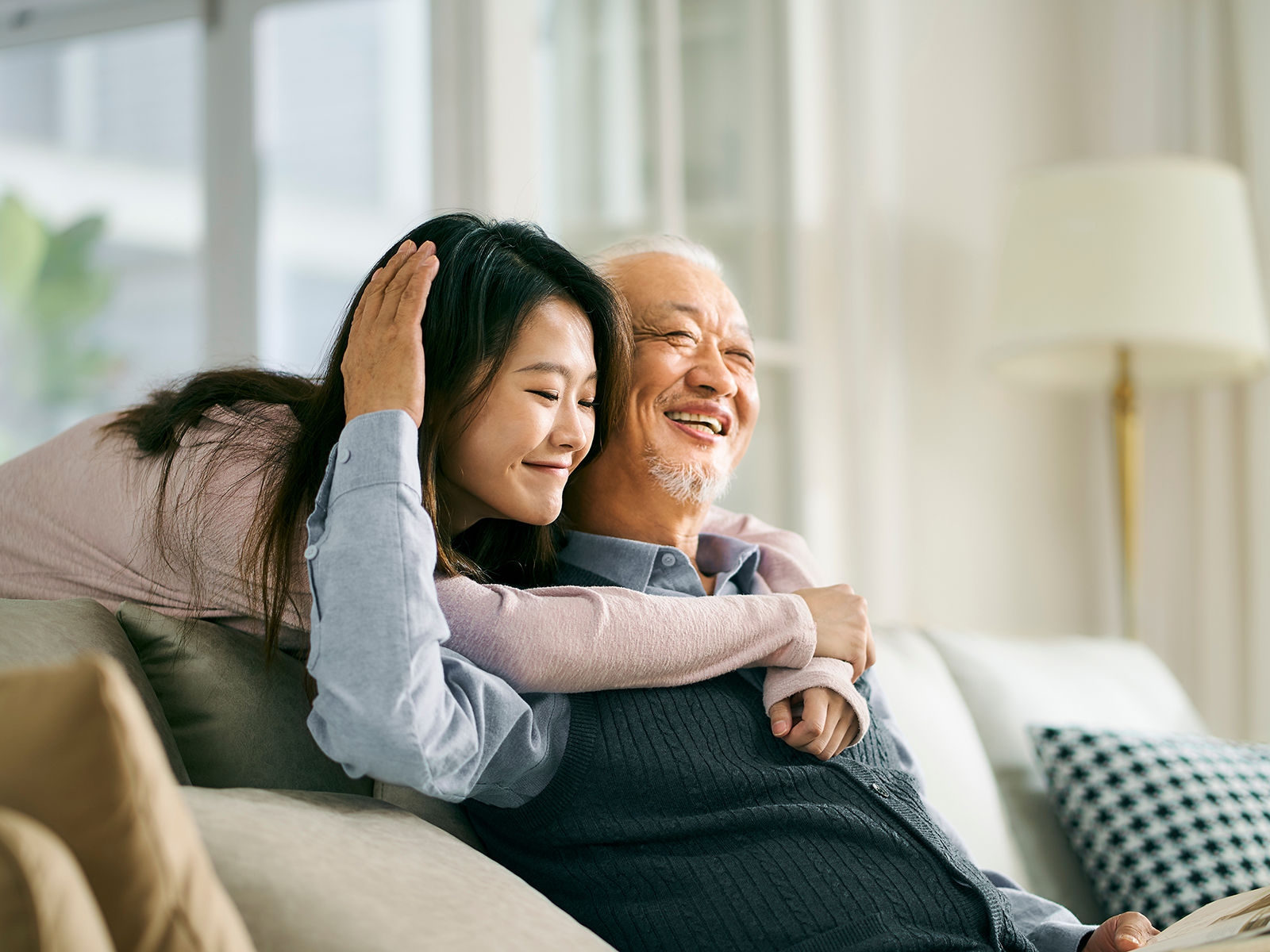 Smiling woman wraps her arms around her father who is sitting on a sofa.