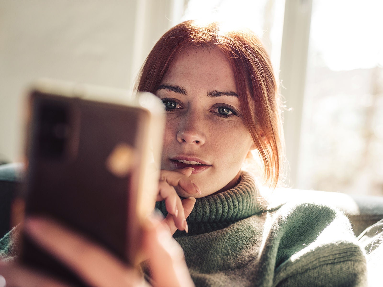 A woman pondering while looking closely at the smartphone held in her hand.