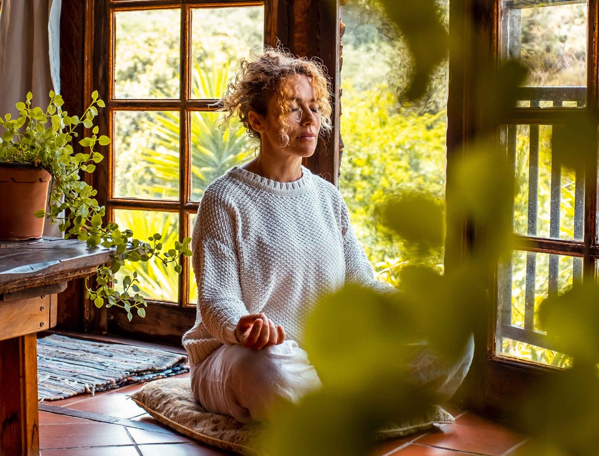 Femme en méditation assise sur un coussin dans une pièce lumineuse.