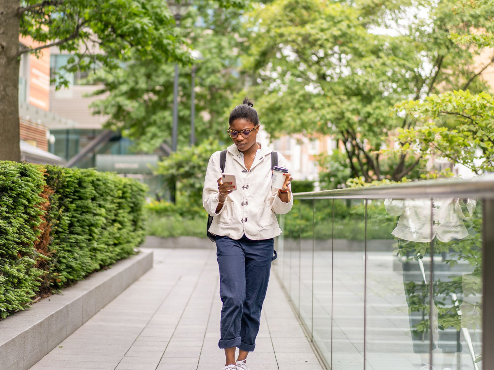 A student walking outside on a school campus, holding a coffee cup in one hand, and looking at her cellphone.