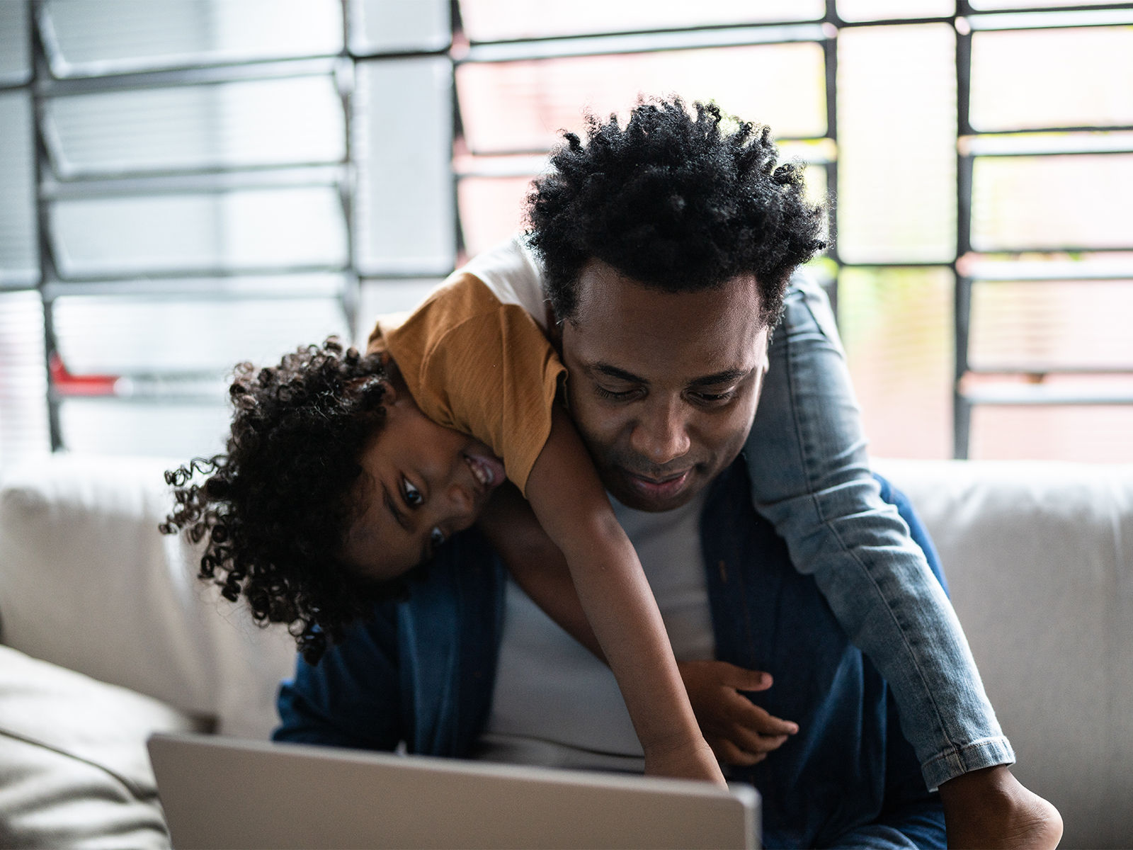 Un père qui regarde son ordinateur portable alors que son enfant souriant est étendu sur ses épaules.