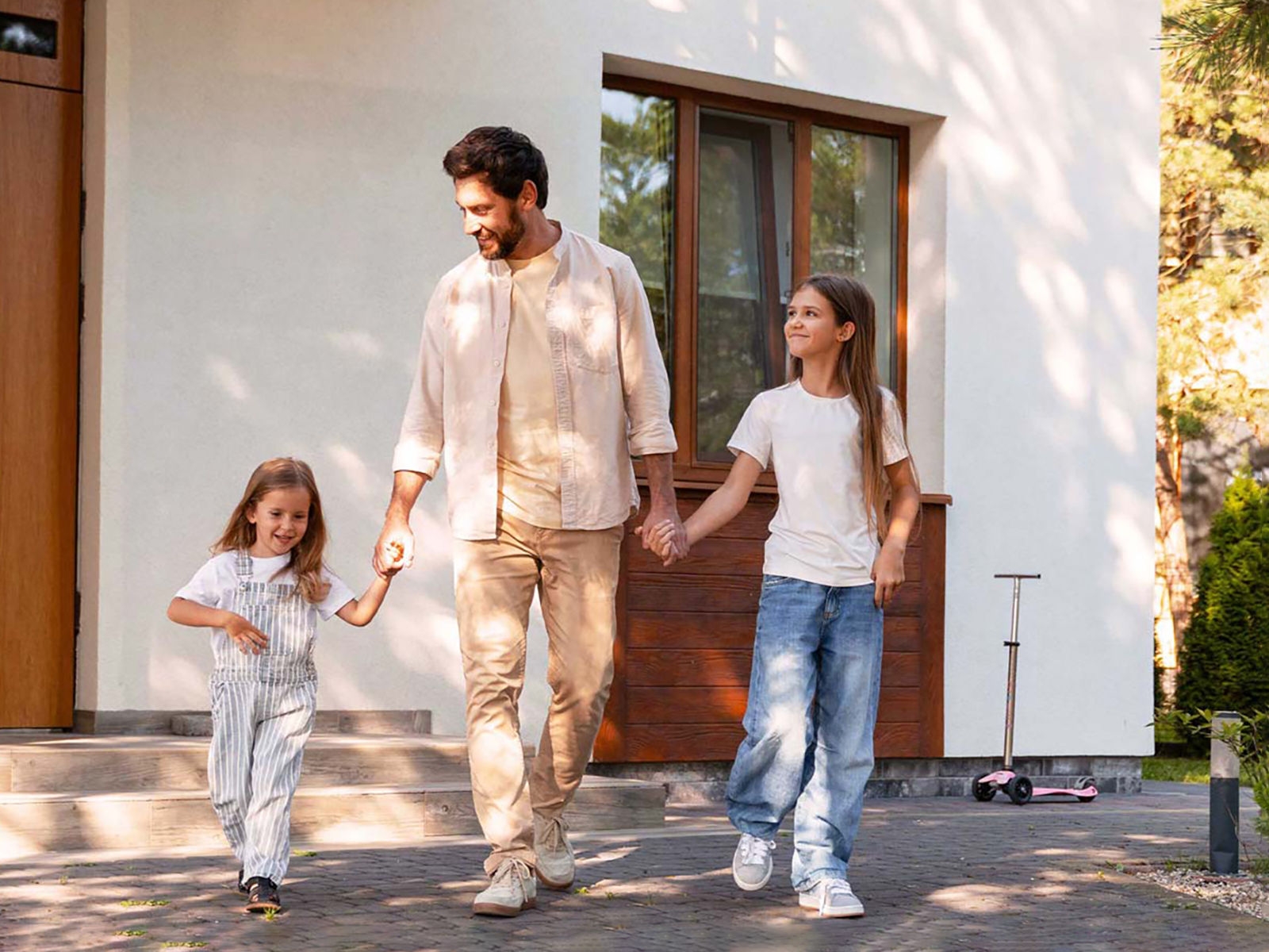 Un homme et ses deux filles souriantes se tiennent la main devant une maison blanche moderne.