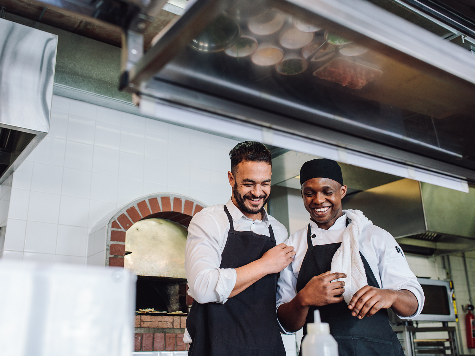 Two smiling men wearing aprons in a restaurant kitchen.