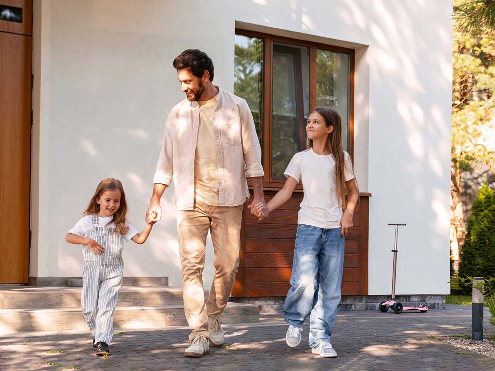 Un homme et ses deux filles souriantes se tiennent la main devant une maison blanche moderne.