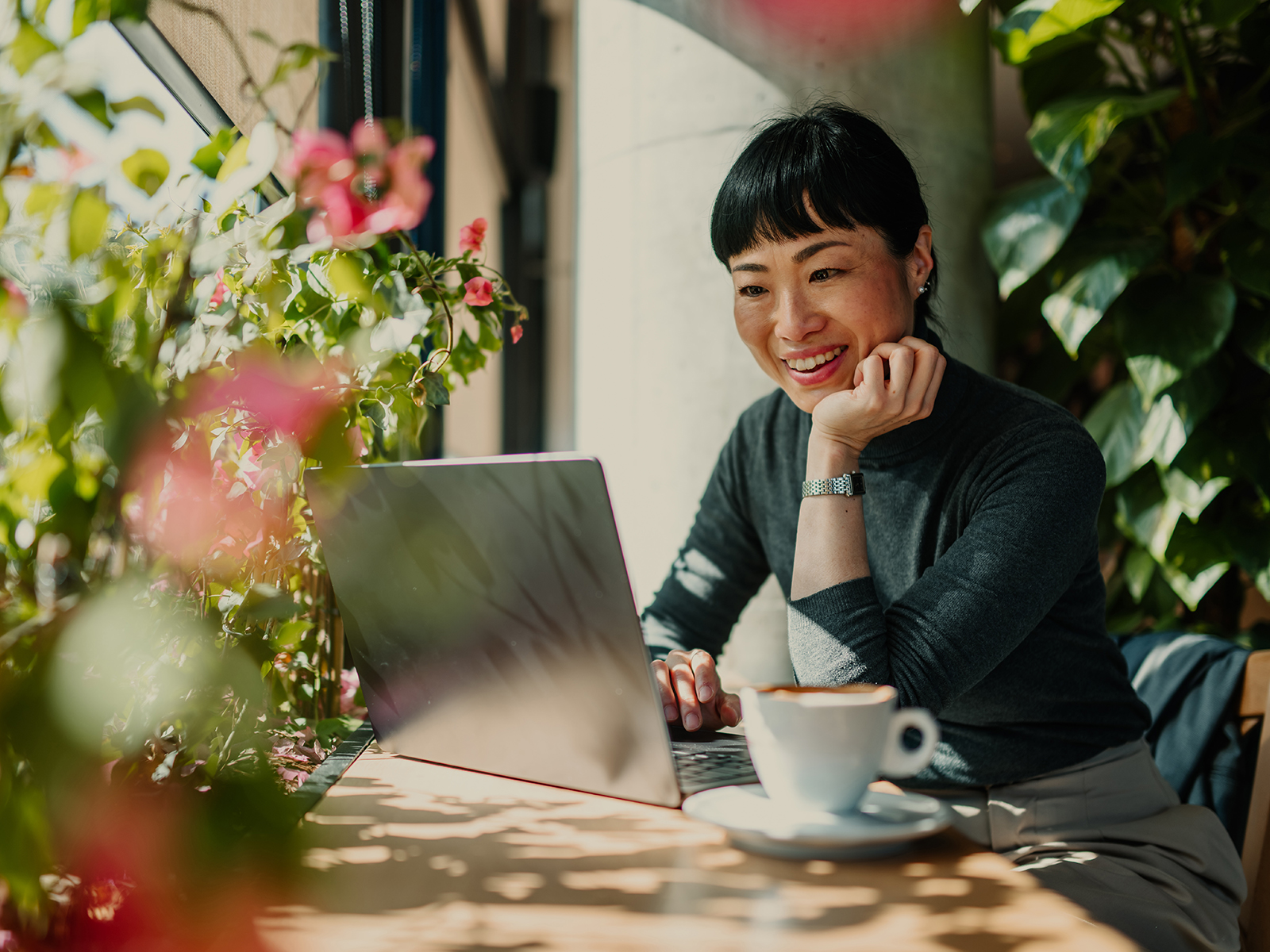 Businesswoman working on laptop in a sunny cafe, enjoying coffee and plants. 