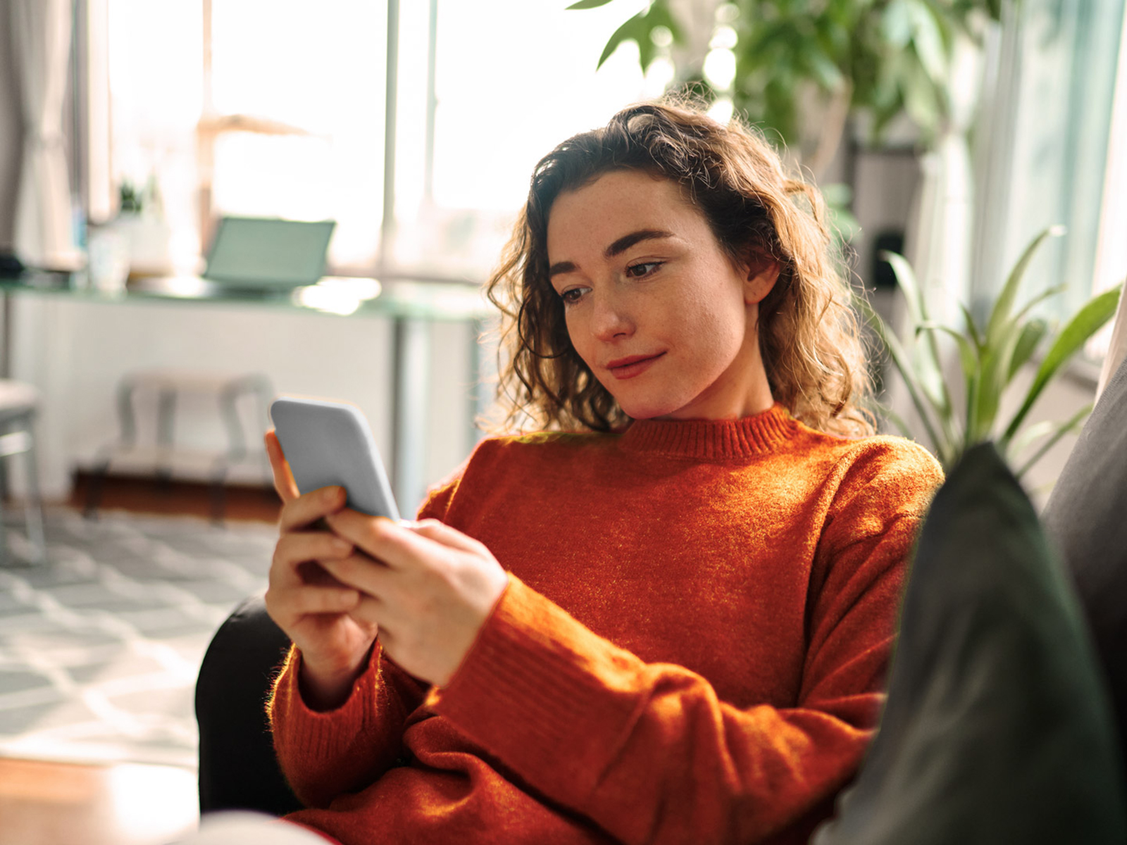 Jeune femme détendue, vêtue d’un pull orange, assise sur un canapé et utilisant un téléphone portable.