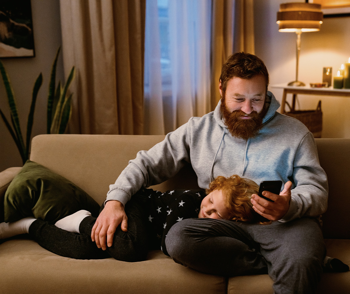 A father sitting on a couch with a young child asleep on his lap.