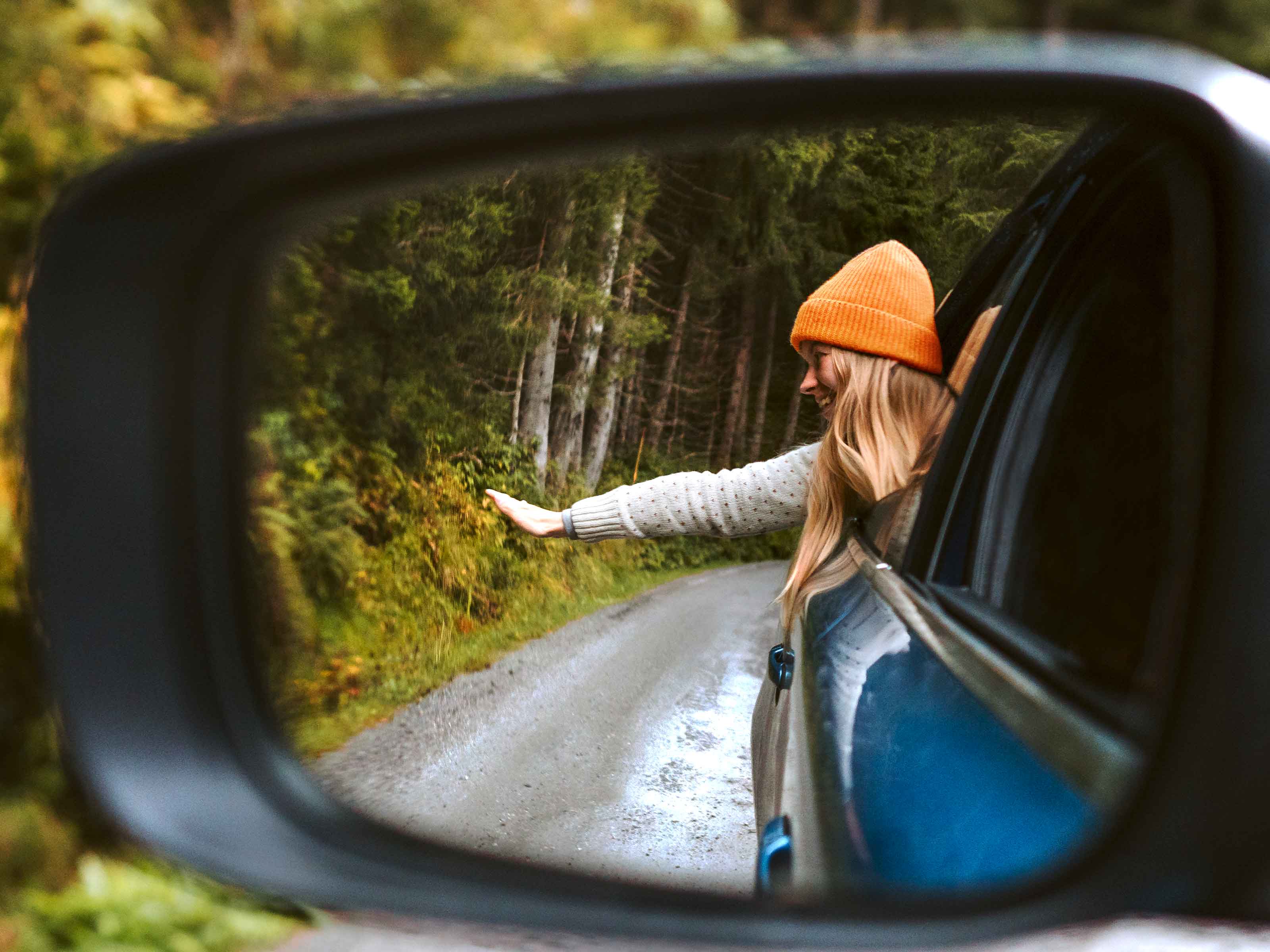 Une femme souriante tend la main par la fenêtre d'une voiture qui roule sur une route de campagne.